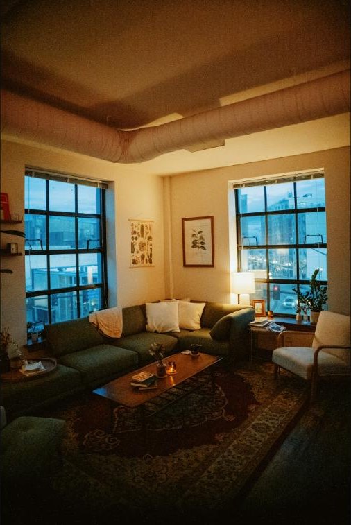 A warm, quiet living room at dusk, a single lamp glowing softly in the corner as evening light fades in the windows